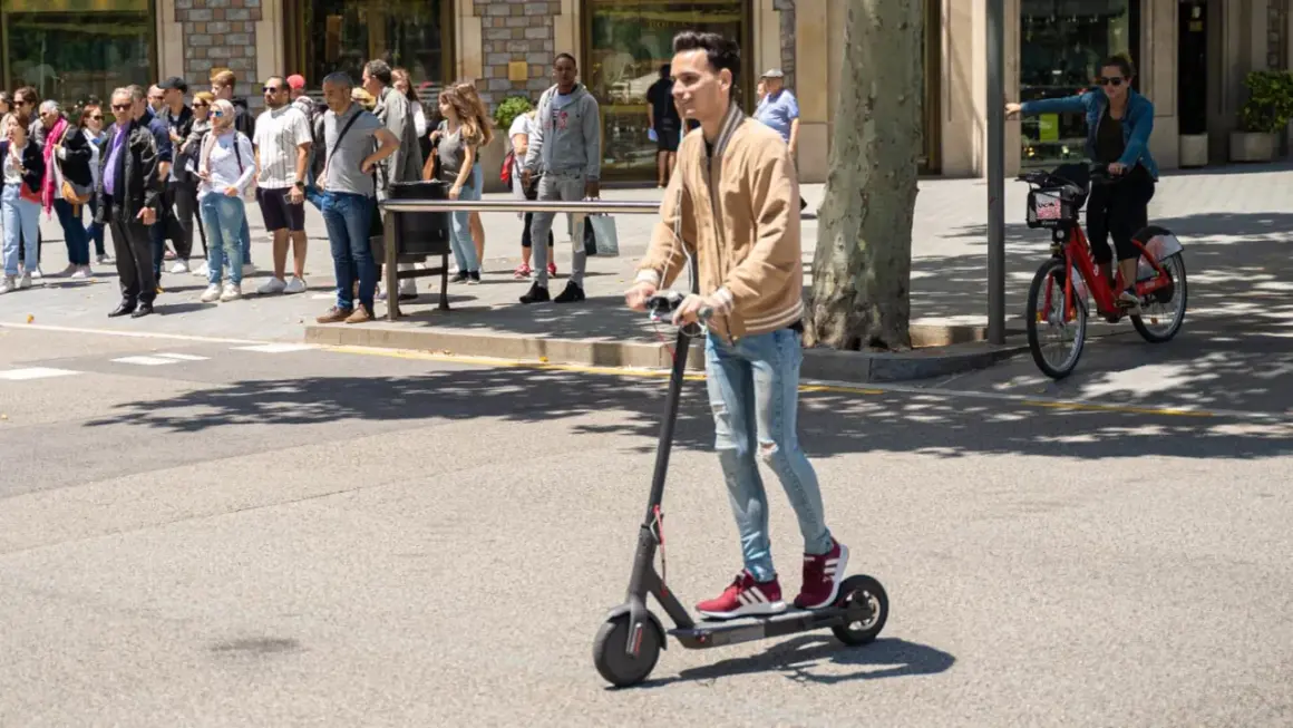 Ein junger Mann fährt mit einem E-Scooter im Ausland. Im Hintergrund warten Menschen auf den Bus und eine Frau fährt mit einem roten Leihfahrrad.