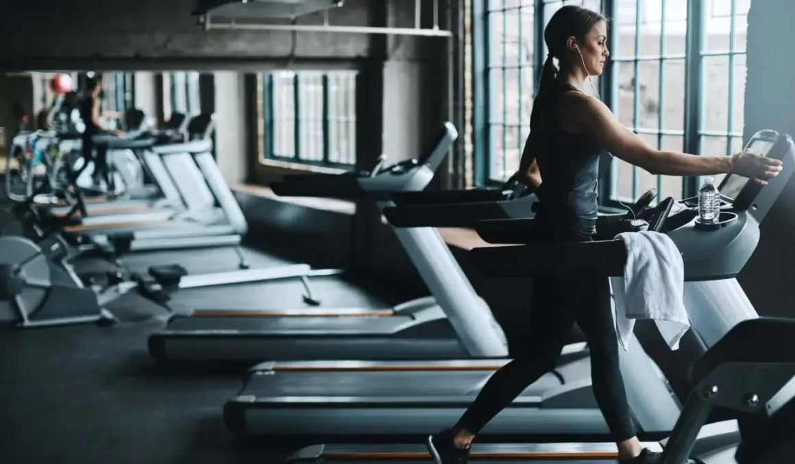Frau im Fitnessstudio beim Laufband Training, mit Kopfhörern und Wasserflasche.