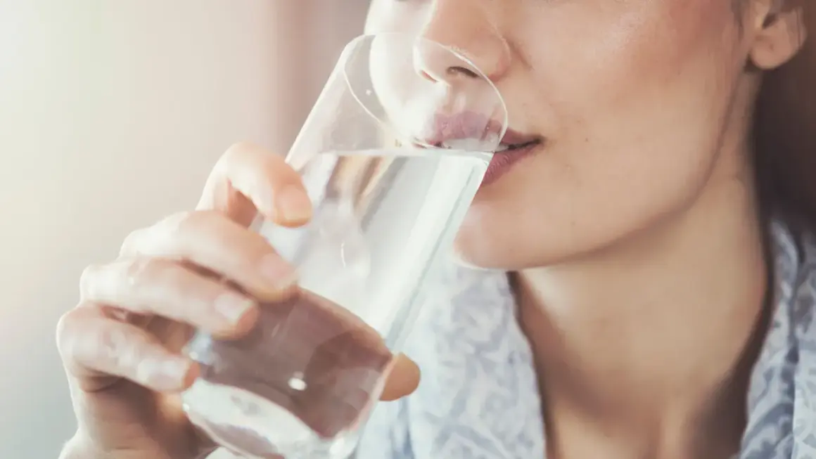 Frau trinkt nach dem Joggen Wasser. Sie nimmt das Glas mit beiden Händen und führt es zum Mund.