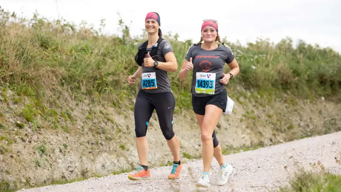 Zwei Läuferinnen auf einer Schotterpiste, die die Halbmarathon Distanz meistern. Sie lächeln und geben Daumen hoch, bereit für die Herausforderung.