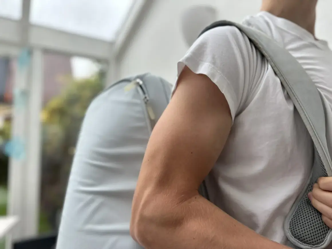 Mann mit Rucksack, der beim Anziehen der Jacke **Schmerzen im Oberarm** spürt.