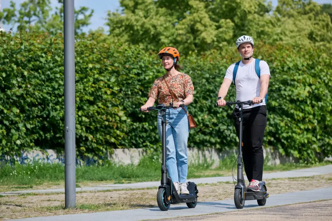 Mann und Frau fahren E-Scooter nebeneinander. Der Unterschied E-Roller und E-Scooter ist hier nicht ersichtlich, beide sind auf dem Weg.