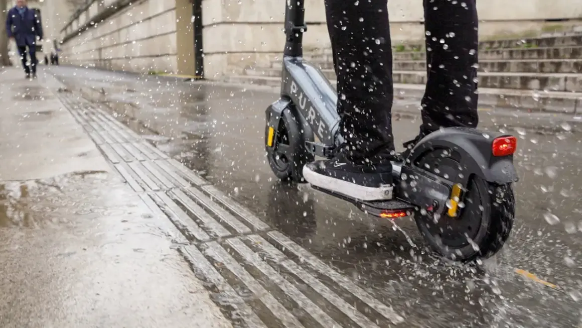Ein e-scooter bei regen spritzt durch Pfützen auf dem nassen Gehweg.