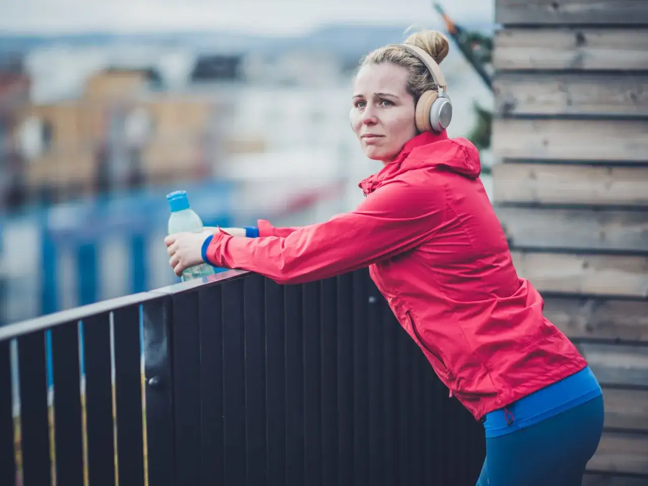 Frau mit Kopfh&ouml;rern und Wasserflasche lehnt an einem Gel&auml;nder. Sie hat leichte Halsschmerzen, aber macht trotzdem Sport.