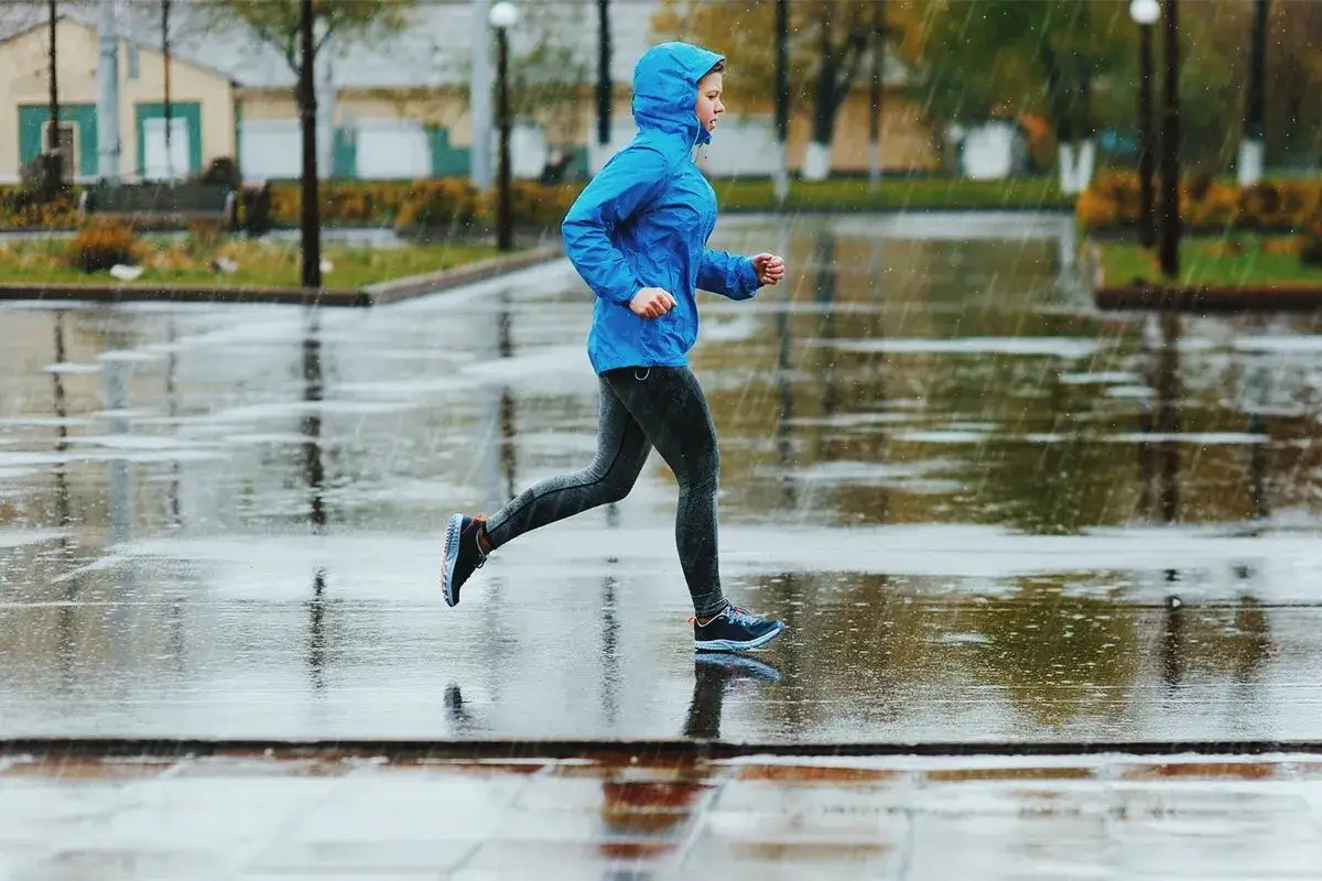 Eine Person in blauer Regenjacke joggt entschlossen durch den Regen auf nassen Pflastersteinen.