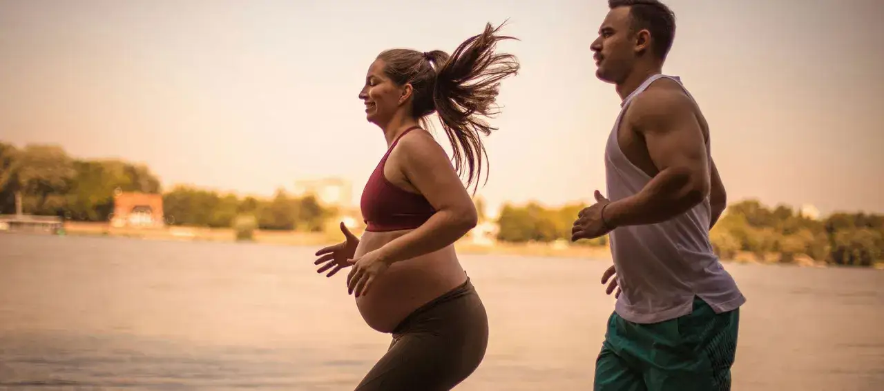 Ein Paar joggt am Wasser entlang. Die Frau ist schwanger und l&auml;chelt gl&uuml;cklich.