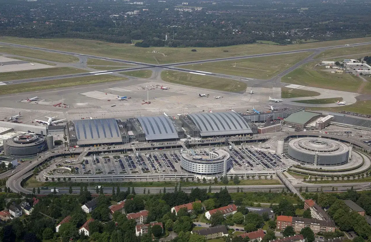 Luftaufnahme des Flughafens Hamburg mit Terminals, Parkhäusern und Rollfeldern. Ein Flugzeug startet, als ob es bei einem airport race Hamburg wäre.