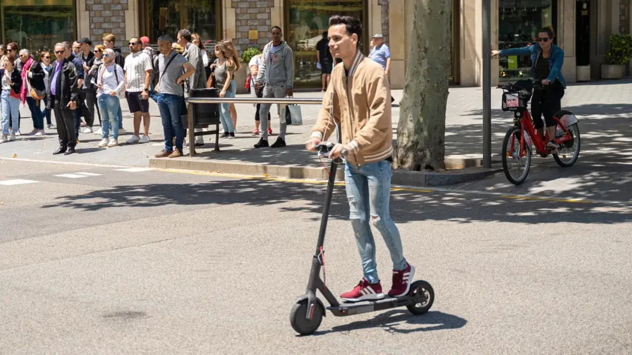 Ein junger Mann fährt mit einem E-Scooter im Ausland. Im Hintergrund warten Menschen auf den Bus.