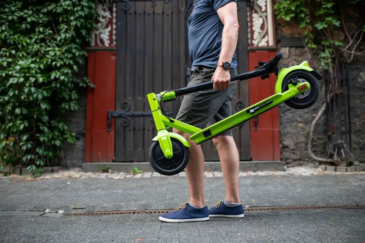 Mann h&auml;lt einen zusammengeklappten E-Scooter in leuchtendem Gr&uuml;n.