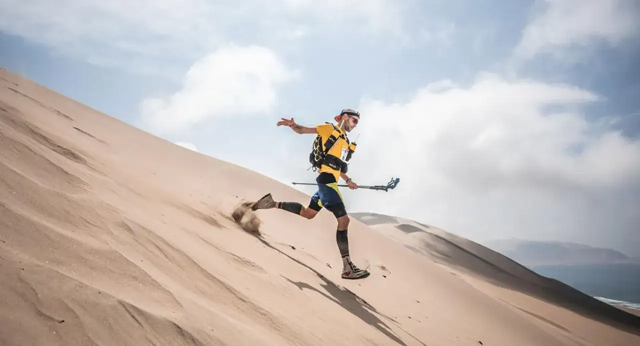 L&auml;ufer st&uuml;rmt eine Sandd&uuml;ne hinunter, Staub wirbelt auf. Ein Teilnehmer des Marathon des Sables k&auml;mpft sich durch die W&uuml;ste.
