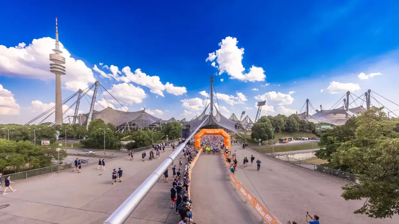 Startschuss für den b2run Munich im Olympiapark. Läufer drängen sich unter dem orangefarbenen Torbogen, im Hintergrund der Olympiaturm und die Zeltdach-Stadien.