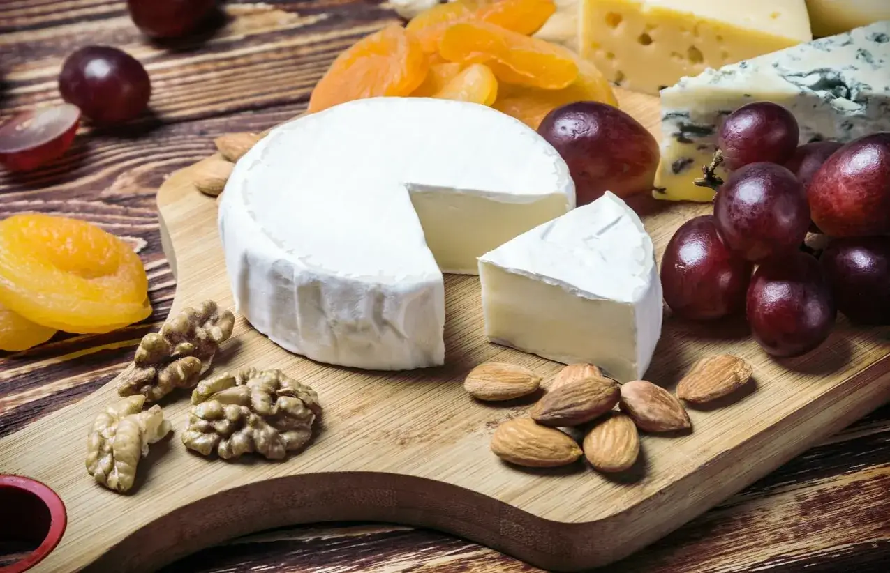 Ein Camembert-K&auml;se mit einem St&uuml;ck, umgeben von Trauben, N&uuml;ssen und Trockenfr&uuml;chten. Ein gesunder Genuss f&uuml;r jeden Anlass.