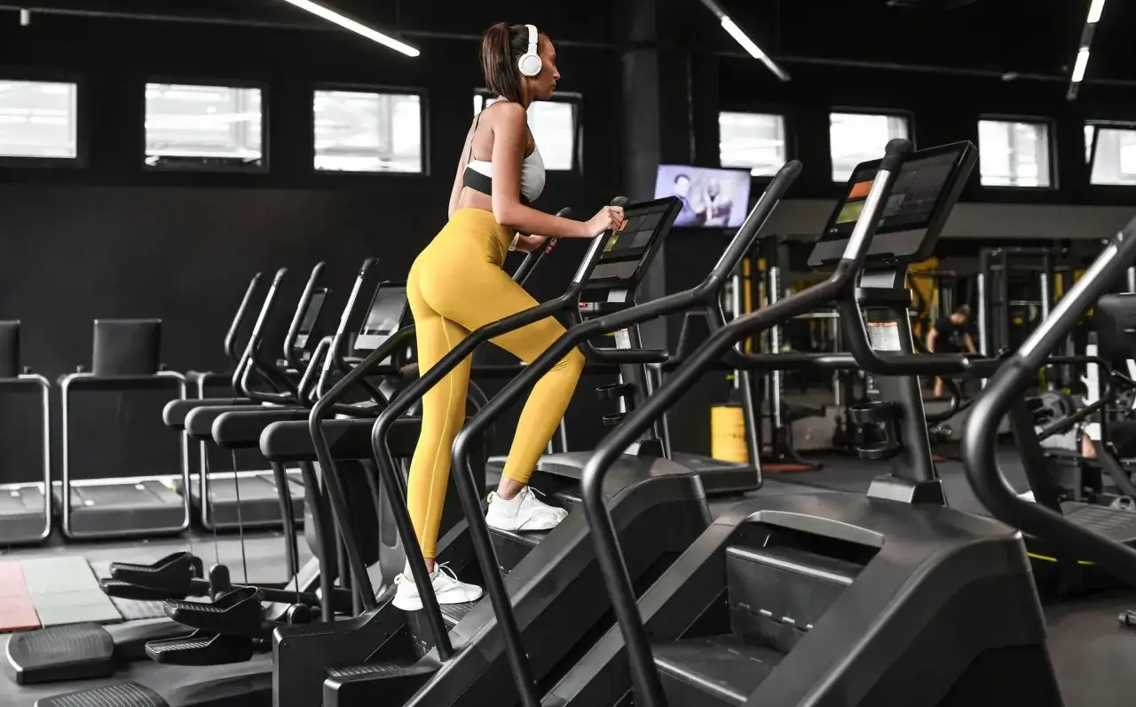 Frau macht ein Stairmaster Workout im Fitnessstudio, trägt Kopfhörer und gelbe Leggings.