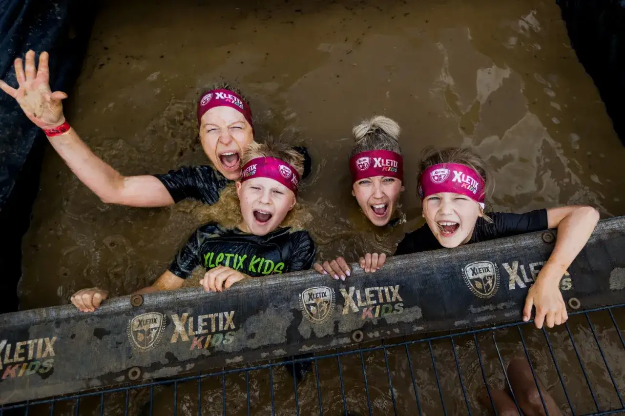 Vier lachende Kinder mit Stirnb&auml;ndern von XLETIX KIDS stecken bis zum Hals in Matsch. Ein unvergessliches Erlebnis beim XLETIX Dresden!
