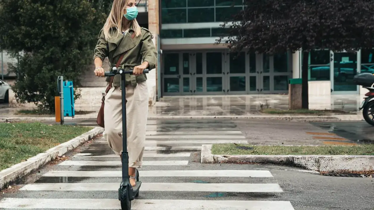 Frau mit Maske f&auml;hrt e scooter bei Regen &uuml;ber einen Zebrastreifen.