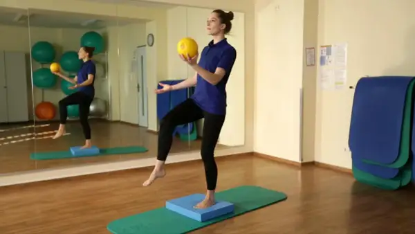 Eine Frau steht auf einem Bein auf einem blauen Kissen, während sie einen gelben Ball hält. Sie übt Balance.