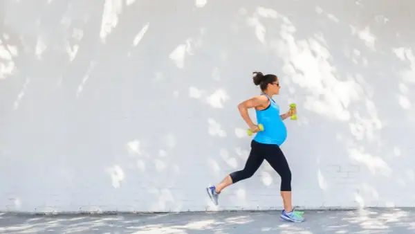 Eine schwangere Frau joggt mit Hanteln vor einer weißen Ziegelwand, beschattet von Baumblättern.