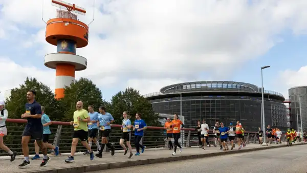 Läufer bei der Airport Race Hamburg, vorbei an einem markanten orange-weißen Turm und einem modernen Gebäude.