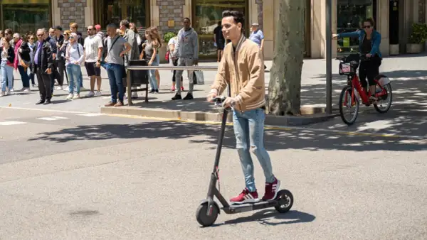 Ein junger Mann fährt mit einem E-Scooter im Ausland. Im Hintergrund warten Menschen auf den Bus und eine Frau fährt mit einem roten Leihfahrrad.