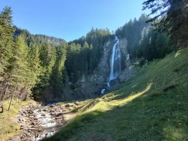Wildstrubel Wasserfall: Ein majestätischer Anblick auf der UTMB-Route. Ein rauschender Wasserfall stürzt über Felsen in ein steiniges Flussbett, umgeben von dichten Nadelwäldern.