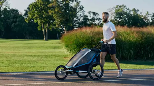 Ein Mann joggt mit Kinderwagen durch eine grüne Parklandschaft.