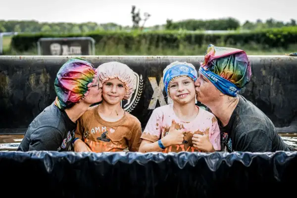 Zwei Erwachsene küssen zwei Kinder auf die Wange, alle tragen bunte Duschhauben. Ein fröhlicher Moment beim XLETIX Dresden Hindernislauf.
